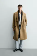 Abrigo largo color camel, de corte relajado (relaxed fit), confeccionado en tejido con mezcla de lana. Presenta cuello con solapas de muesca, manga larga, bolsillos de vivo en cadera, bolsillo interior y cierre frontal de botonadura cruzada.