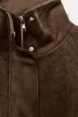 Chaqueta de tejido efecto ante color marrón, con cuello alto, cierre frontal con cremallera y botones a presión, manga larga y bolsillos de plastrón con solapa en el delantero.