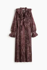 Vestido largo de manga globo, color marrón oscuro, con estampado paisley en tonos claros. Presenta cuello alto con volados y escote en V con lazos ajustables, y puños con volados.