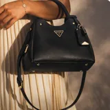 Cartera Guess modelo Meridian II, de diseño estructurado en color negro con acabado texturizado. Presenta doble asa de mano, correa ajustable y extraíble, y detalle de logo triangular metálico en el frente junto con un charm colgante.