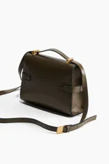 Cartera bandolera rectangular color verde oliva, de cuero sintético, con correa fina ajustable y solapa con cierre magnético. Presenta una trabilla decorativa con hebillas doradas en la parte delantera.