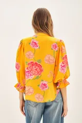 Camisa de viscosa con estampado floral en tonos amarillos y rosas. Presenta escote redondo con cierre frontal de botones, mangas al codo con terminación elástica y detalle de volado.