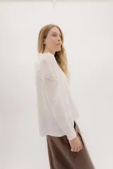 Camisa de lino color beige con mangas abullonadas y detalles de puntilla negra en forma de V en el frente. Presenta cuello a la base con borde desflecado, cierre frontal con botones forrados y puños con alforzas.