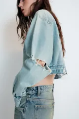 Cazadora corta de denim en tono celeste claro, con cuello solapa, manga larga y cierre frontal con botones. Presenta un bolsillo de plastrón en el delantero y detalles de rotos en el cuello y el ruedo deshilachado.