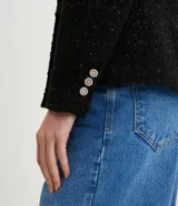 Blazer negro de tweed con hilos metalizados, corte entallado, escote en V profundo, bolsillos delanteros con solapa y doble botonadura con seis botones decorativos de strass.