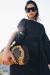 Bolso tipo pouch confeccionado completamente con cuentas de madera en tonos marrón, beige y tostado, formando un patrón abstracto. Cuenta con una correa fina de cadena dorada desmontable y cierre superior con cremallera.