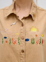 Camisa sin mangas de denim ligero, color celeste lavado, con cuello camisero y botones frontales. Presenta bordados temáticos de playa/mar en el pecho, incluyendo sol, olas, gaviotas, peces y corales/vegetación marina.
