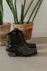 Botinetas de cuero negro con diseño estilo western, punta cuadrada y taco bajo con acabado desgastado. Presentan una tira decorativa con estribo metálico desmontable alrededor del tobillo.