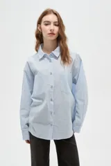 Camisa de algodón a rayas verticales en tonos celeste y blanco, con cuello camisero, cierre frontal de botones y dos bolsillos delanteros tipo parche. Presenta un corte holgado y mangas largas.