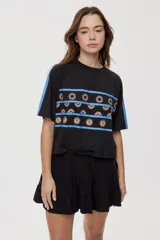 Remera negra de manga corta con calce relajado y cuello redondo. Presenta franjas azules en los hombros y mangas, y un diseño estampado frontal con franjas horizontales azules y motivos gráficos.