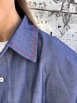 Sobre camisa de gabardina símil denim, color azul, con costuras visibles en color rojo y etiqueta de cuero en la espalda. Presenta un corte holgado y mangas largas.