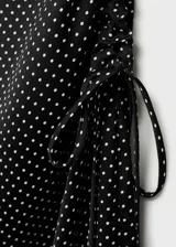Vestido midi de tejido fluido con estampado de lunares blancos sobre fondo negro. Presenta un diseño entallado sin mangas con escote redondo, tirantes anchos y un detalle de fruncido lateral con lazo que crea un efecto drapeado. Incluye cierre de cremallera invisible en la parte posterior.