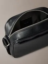 Cartera tipo cámara Calvin Klein de color negro, confeccionada en poliuretano texturizado. Presenta una silueta compacta con cierre de cremallera superior, correa de hombro ajustable con el logo de la marca y monograma CK grabado en la esquina frontal.