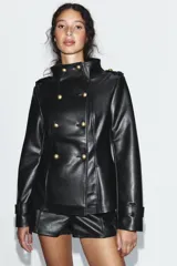 Chaqueta entallada de efecto piel en color negro, con cuello subido y cierre frontal cruzado con botones metálicos dorados. Cuenta con manga larga con trabillas en los puños y hombreras decorativas.