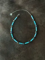 Collar corto de diseño artesanal compuesto por cuentas cilíndricas de vidrio en tono celeste y pequeñas piedras naturales esféricas en tonos azulados, intercaladas con detalles metálicos. Incluye cadena de acero quirúrgico para ajuste de largo.