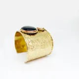 Brazalete ancho de bronce con acabado martillado, que presenta dos piedras circulares engarzadas: una de nácar blanco de gran tamaño y otra de piedra natural oscura de menor tamaño. Es una pieza de diseño ajustable tipo cuff.