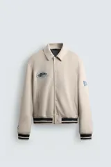 Cazadora estilo varsity de tejido efecto ante en color beige. Presenta cuello solapa, cierre frontal con botones a presión y puños y ruedo con acabado en rib a rayas negras. Incluye parches bordados decorativos en el pecho y la manga, además de bolsillos laterales de doble vivo.