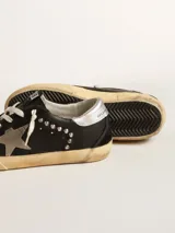 Zapatillas Golden Goose modelo Super-Star, confeccionadas en cuero negro con detalles en gamuza, estrella lateral gris y tachas metálicas plateadas. Cuentan con suela de goma con acabado envejecido y cordones de algodón crudo.
