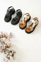 Sandalias negras de estilo chunky con plataforma dentada. Presentan tiras ajustables con cierre de velcro y hebillas metálicas, combinando texturas de cuero granulado y detalles en charol.