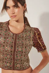 Saco corto estilo crop top con cuello redondo y mangas cortas. Confeccionado en tejido jacquard con lurex, presenta un estampado en tonos borgoña y dorado, y está ricamente bordado a mano con hilos y espejos (mostacillas). Cierre frontal con ganchos ocultos.