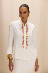Camisa de color blanco hueso con cuello clásico, mangas tres cuartos y corte recto. Presenta un aplique decorativo frontal compuesto por piedras de colores y detalles metálicos dorados.