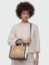 Cartera tipo tote bag Marc Jacobs en color tostado, con detalles en negro y blanco. Cuenta con las inscripciones "THE TOTE BAG" y "MARC JACOBS" estampadas en el frente. Posee doble asa de mano superior y correa de hombro ajustable y desmontable.