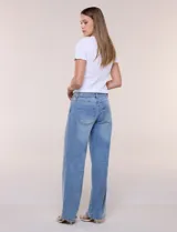 Jean wide leg de tiro medio, color azul claro, confeccionado en denim de algodón con un toque de elastano. Cuenta con cinco bolsillos, presillas para cinturón y cierre frontal con botón. El diseño destaca por su pretina regulable con cruce y broches metálicos.