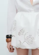 Falda corta blanca de estilo globo, con cintura elástica y diseño abullonado en el bajo. Presenta delicados detalles florales bordados.