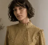 Blusa color mostaza con textura de pequeños puntos y detalles calados verticales. Presenta cuello alto con volados, volados decorativos en los hombros y mangas largas con puños elastizados.