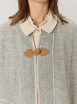 Capa de tejido de punto en color gris melange, con diseño abierto y cierre frontal mediante un botón tipo duffle de madera con detalle en cuero. Presenta bolsillos laterales amplios y bordes con costuras a la vista.
