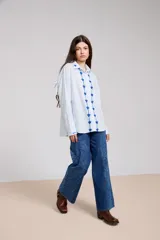 Camisa de corte holgado color celeste claro, con bordados florales azules a lo largo de la botonadura frontal y en el cuello. Presenta mangas largas con detalle de lazo en los hombros y puños abotonados.