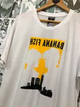 Remera blanca de manga corta con estampado de siluetas de personajes del anime Banana Fish sobre un fondo amarillo y un skyline negro.