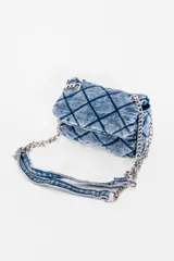 Cartera bandolera de jean negro con diseño acolchado en forma de rombos. Tiene correa de cadena plateada y manija de jean.
