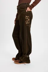 Jean tipo baggy de pierna ancha y recta, color marrón oscuro. Presenta detalles de costuras decorativas, bolsillos laterales redondeados y bolsillos traseros grandes. Incluye tiras ajustables con hebillas metálicas en los tobillos para un efecto fruncido.