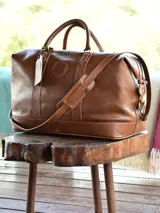 Bolso de viaje marrón de cuero liso con correa al hombro ajustable y extraíble. Tiene manijas superiores reforzadas, cierre superior con cremallera y etiqueta de cuero colgante.