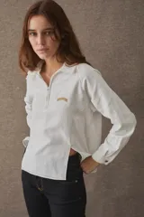 T-shirt de manga larga confeccionada en mezcla de algodón y lino, color blanco, con cuello camisero, cierre frontal parcial con cremallera, puños abotonados y aberturas laterales en el bajo. Cuenta con un pequeño bordado distintivo en el pecho.