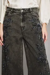 Pantalon de denim de corte amplio y tiro medio, color gris oscuro con efecto lavado. Presenta bordados decorativos con pedrería a lo largo de los laterales de las piernas.