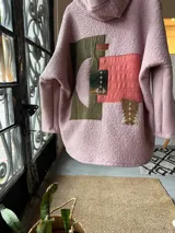 Campera tipo hoodie oversize de corderito rosa, con diseño de collage en la espalda que combina parches de tela en tonos verde oliva, rosa y estampados geométricos étnicos.