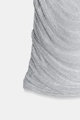 Vestido corto de tejido mesh plateado con aplicación de strass. Presenta cuello redondo, diseño sin mangas con drapeado en los hombros y una superposición efecto capa. Incluye frunces en los laterales y cierre oculto en el cuello.