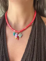Collar corto con cuentas tubulares de cerámica color rojo vibrante, con dijes colgantes de cerámica y cristal checo: hoja turquesa, figura alargada lila con base amarilla, chile rojo y caracola celeste.