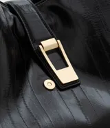 Bolsa tipo shopper de tamaño grande, confeccionada en poliuretano texturizado con acabado brillante. Presenta un diseño estructurado con correas fijas para el hombro, cierre superior mediante botón a presión y un detalle metálico decorativo en la parte frontal.