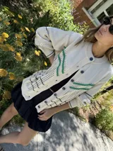 Saco de punto color crema con detalles en crochet verde, cierre frontal con botones y mangas largas.