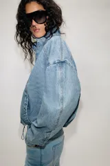 Cazadora bomber de denim con cuello subido, manga larga y cierre frontal con cremallera y botones a presión ocultos. Presenta costuras verticales decorativas y bajo con ajuste elástico.