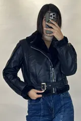 Campera tipo aviador de cuero sintético negro con interior y detalles de piel sintética al tono en cuello, puños y ruedo. Presenta cierre frontal cruzado y cinturón ajustable con hebilla metálica en la cintura.