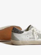 Championes Golden Goose modelo Super-Star, confeccionados en cuero color blanco con paneles de gamuza gris. Presentan el icónico parche de estrella lateral, detalle de glitter en el talón, efecto envejecido en la suela y logo GGDB grabado en el lateral.