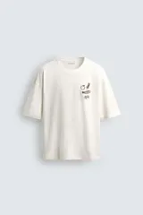 Camiseta de manga corta color blanco roto, corte relajado, con un pequeño bordado/estampado en el pecho que incluye una manzana, una rama de laurel, una trompeta y dos dados.