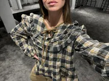 Camisa de manga larga con estampado a cuadros en tonos gris, negro y mostaza. Presenta volados en la parte superior delantera y en los hombros, con un diseño de corte holgado y lazos decorativos en el cuello.