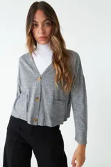 Cardigan de tejido de punto color gris melange, con escote en V, cierre frontal con botones y bolsillos tipo cartera. Presenta un diseño de corte recto y mangas largas.