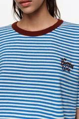 Remera estilo boxy de algodón con diseño de rayas horizontales verdes sobre fondo crudo. Cuenta con cuello redondo marrón tostado, mangas cortas caídas y detalle de logo bordado en el pecho.