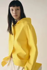 Cazadora impermeable de acabado engomado en color amarillo, con capucha ajustable, cierre frontal con botones a presión, bolsillos laterales y mangas largas ajustables.